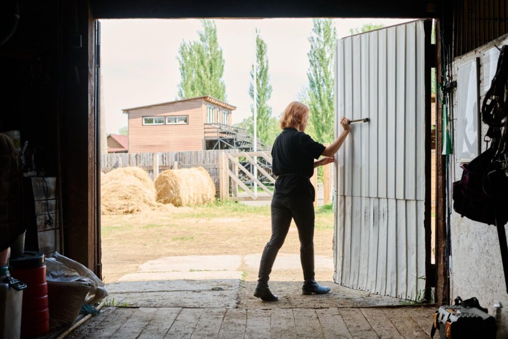 A person opening a large metal barn door, revealing a bright farm scene with hay bales and a farmhouse. Represents functional barn restoration and upgrades.