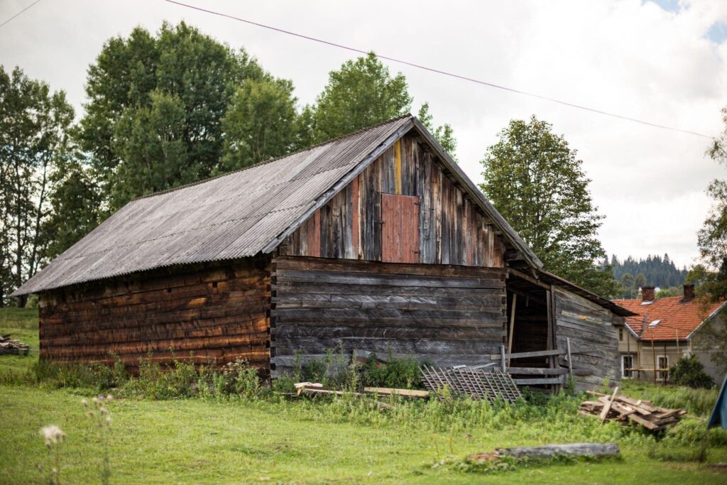 Old wooden barn with weathered siding and a corrugated roof, sitting on a grassy lot—example of a structure needing barn repair or restoration