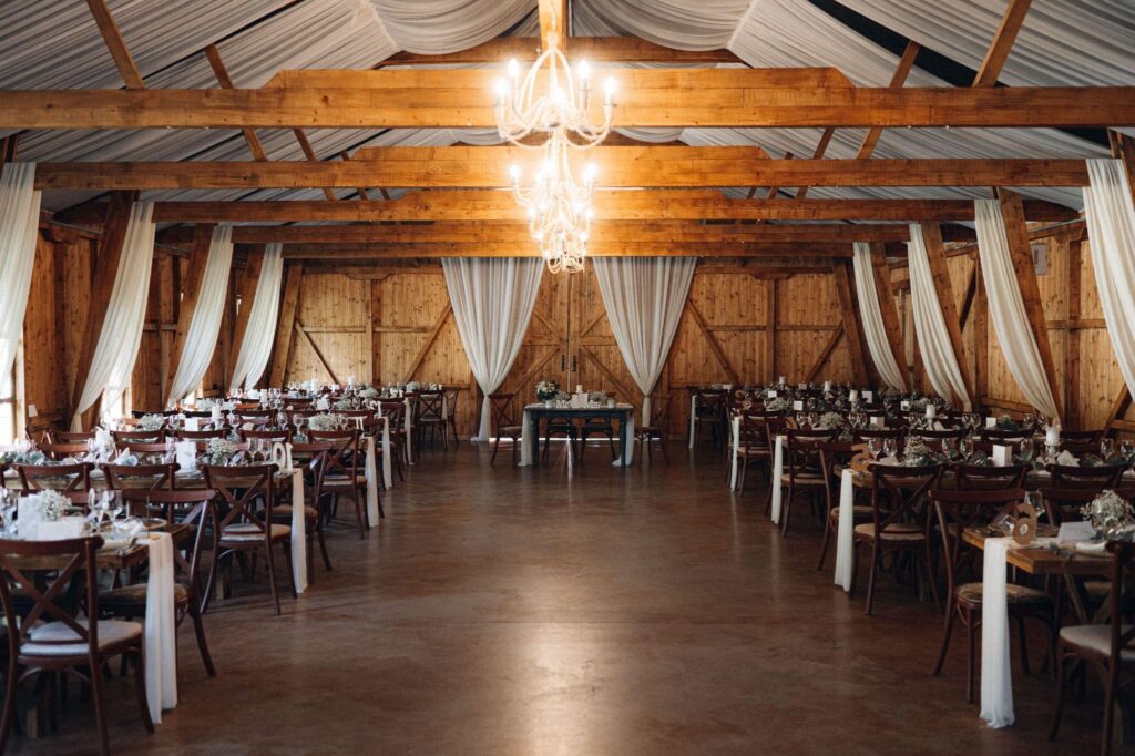 Rustic barn wedding venue with wood beams, chandeliers, and rows of tables. A perfect example of barn construction near you for events.