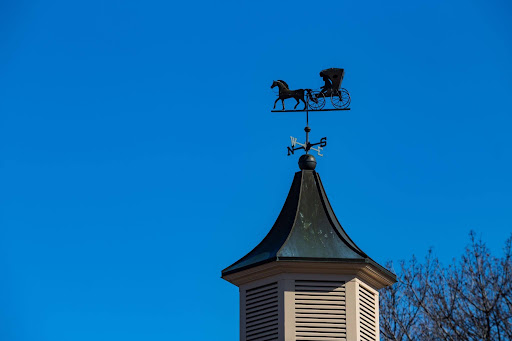 Louvered barn cupola with a horse and carriage weathervane against a clear blue sky